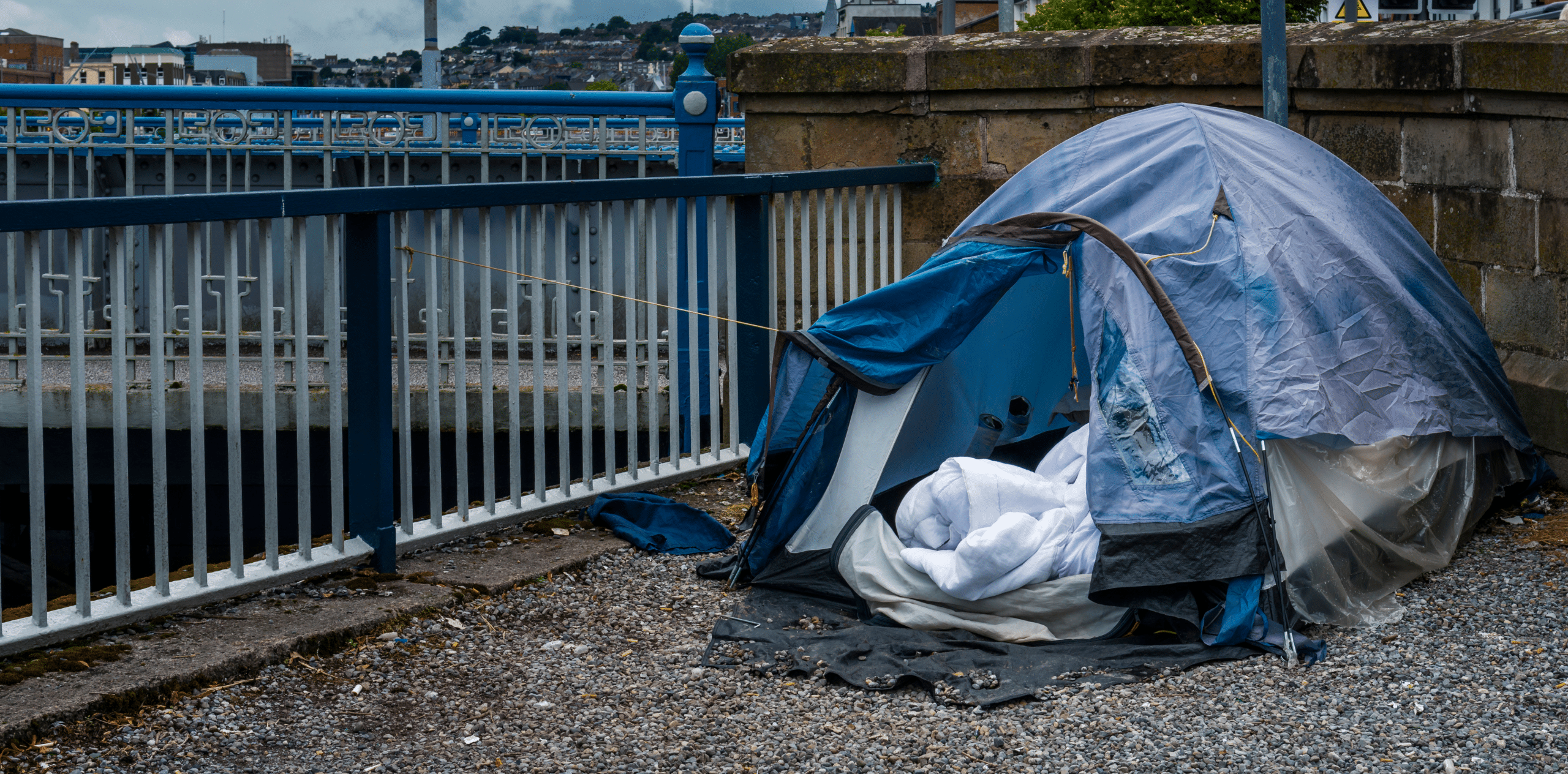Blue camping tent pitched on a gravel surface beside a blue railing and stone wall, with bags and bedding around it in an urban riverside setting.
